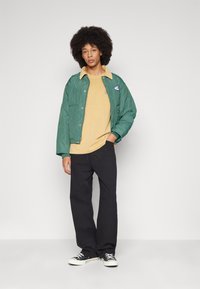 Chaqueta de punto verde, camisa amarilla, pantalones negros y zapatillas negras. La chaqueta presenta un cuello en contraste y un logo en el pecho.