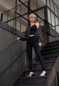 Fiatal személy rövid szőke hajjal, fekete farmert, fekete ujjatlan felsőt és félig elhasználódott farmerdzsekit visel, fém lépcsőkön állva támaszkodik a korlátra.