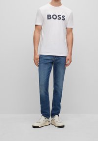 Vit T-shirt med rund hals och stor svart "BOSS" logga, i kombination med blå denimjeans och beige sneakers med svarta detaljer.