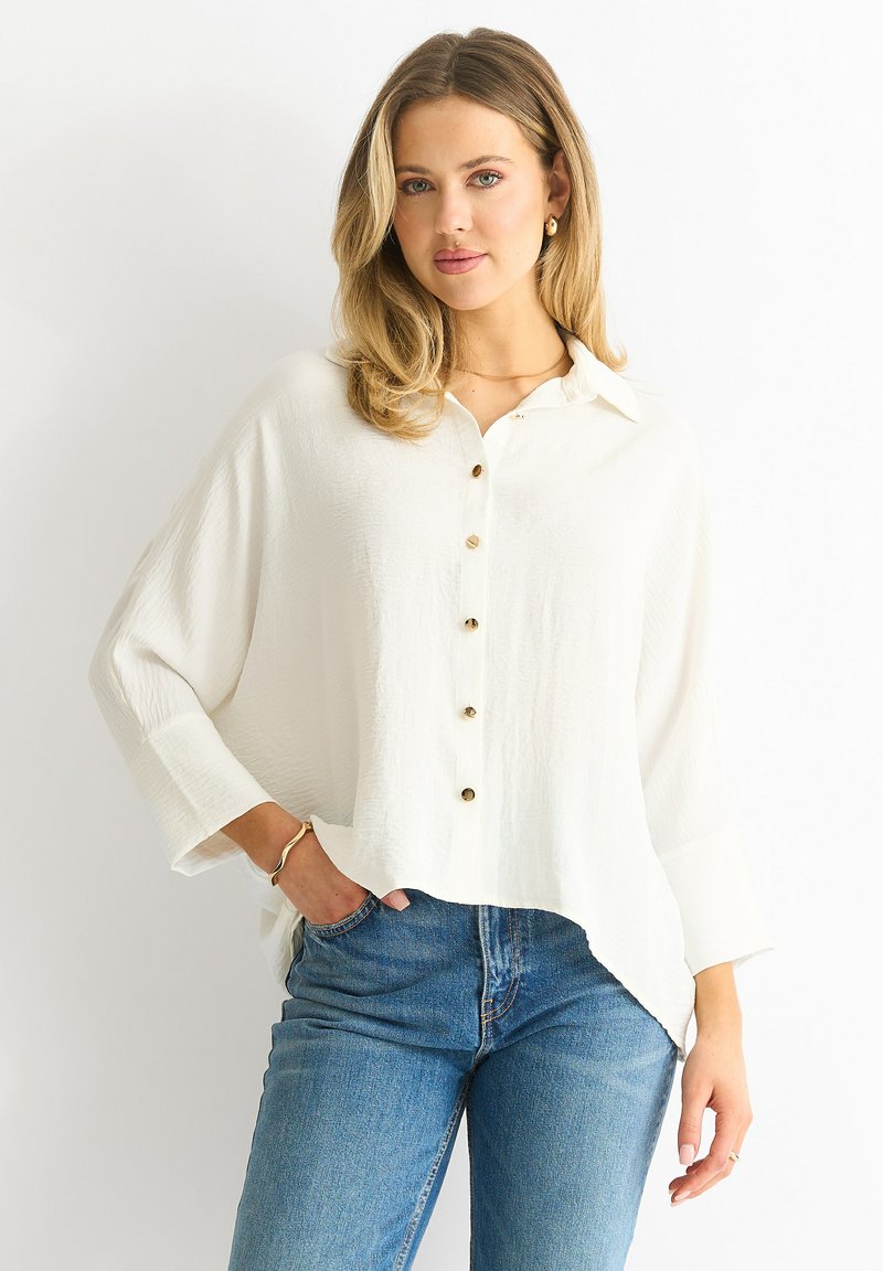 Witte blouse met lange mouwen, gestructureerde stof, ronde zoom en gouden knopen. Gecombineerd met blauwe spijkerbroek.