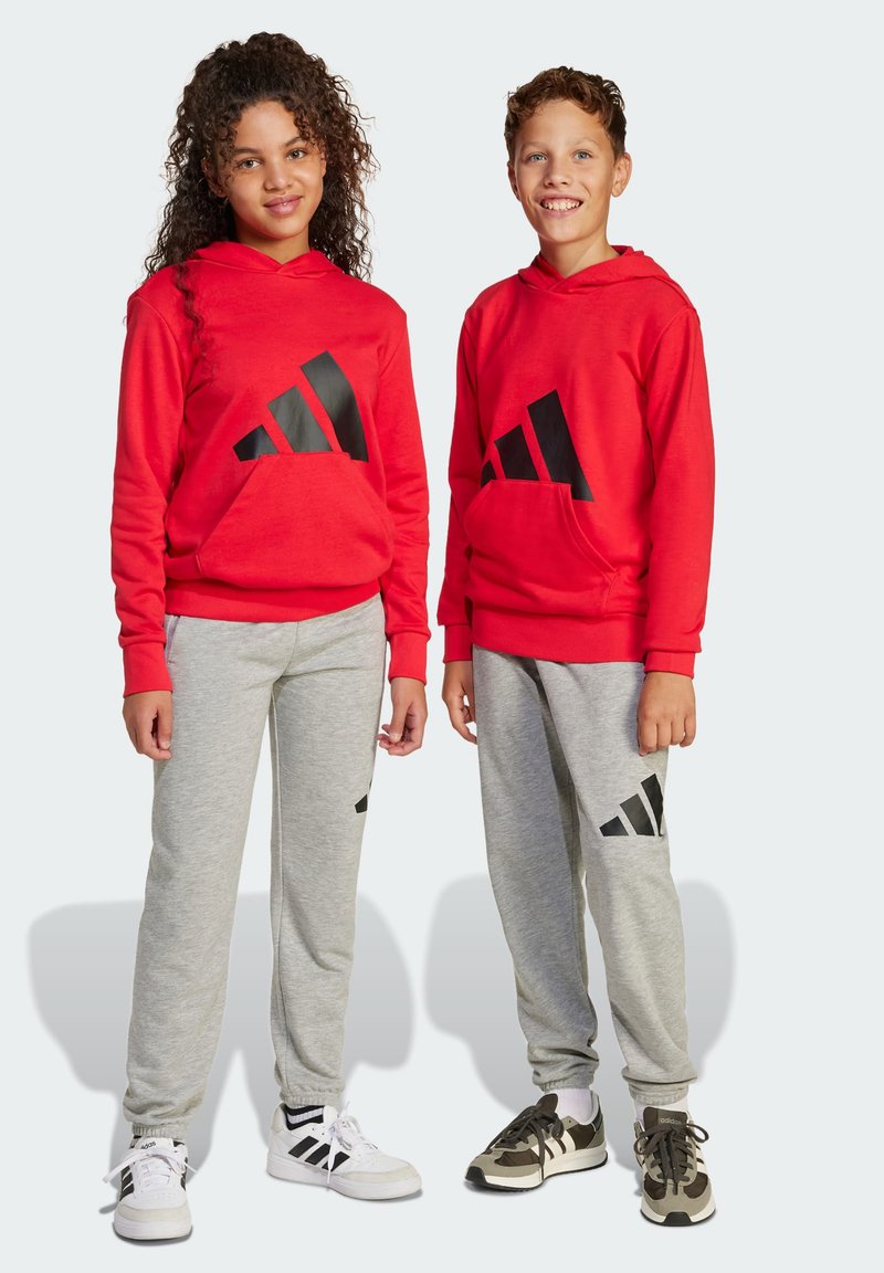 Des hoodies rouges avec logo noir, des pantalons de survêtement gris avec des accents de logo et des baskets. Le tissu semble doux et décontracté, adapté pour une tenue sportive.