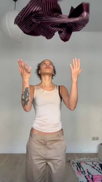 Asmuo su tatuiruote ant kairės rankos metantis į orą bordo spalvos dryžuotą marškinį minimaliame kambaryje su kilimu ir elektros lizdu.