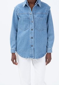 Chemise en denim bleu clair avec un col pointu, deux poches poitrine, un boutonnage à l'avant et des manches longues. Présente un ourlet arrondi et un tissu texturé.