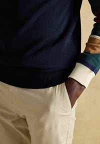 Osoba w beżowych spodniach, trzymająca jedną rękę w kieszeni, w granatowym swetrze oraz wielokolorowej, pasiastiej bluzce z długim rękawem widocznej na nadgarstku.