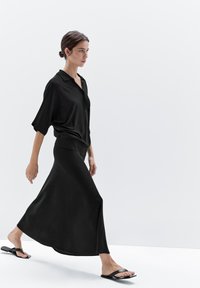 Black flowing skirt and loose black shirt, both made of smooth fabric. Model wears black flat sandals, walking against a plain light background.