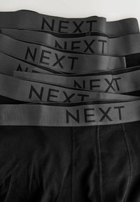 Pila de calzoncillos bóxer negros con bandas grises en la cintura etiquetadas como "NEXT", dispuestos en un patrón superpuesto y en capas.