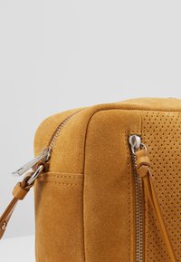 Acercamiento a la esquina de un bolso de ante color beige con cremalleras metálicas, tiradores de cuero y un panel de ante perforado sobre un fondo gris.