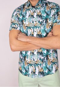 Chemise à manches courtes présentant un motif de feuilles de palmier tropicales en vert, jaune et noir sur un fond blanc ; détail des boutons visible.