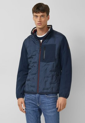 Mężczyzna w granatowej pikowanej kurtce z kieszenią na zamek na piersi i w niebieskich dżinsach, stojący na neutralnym tle.