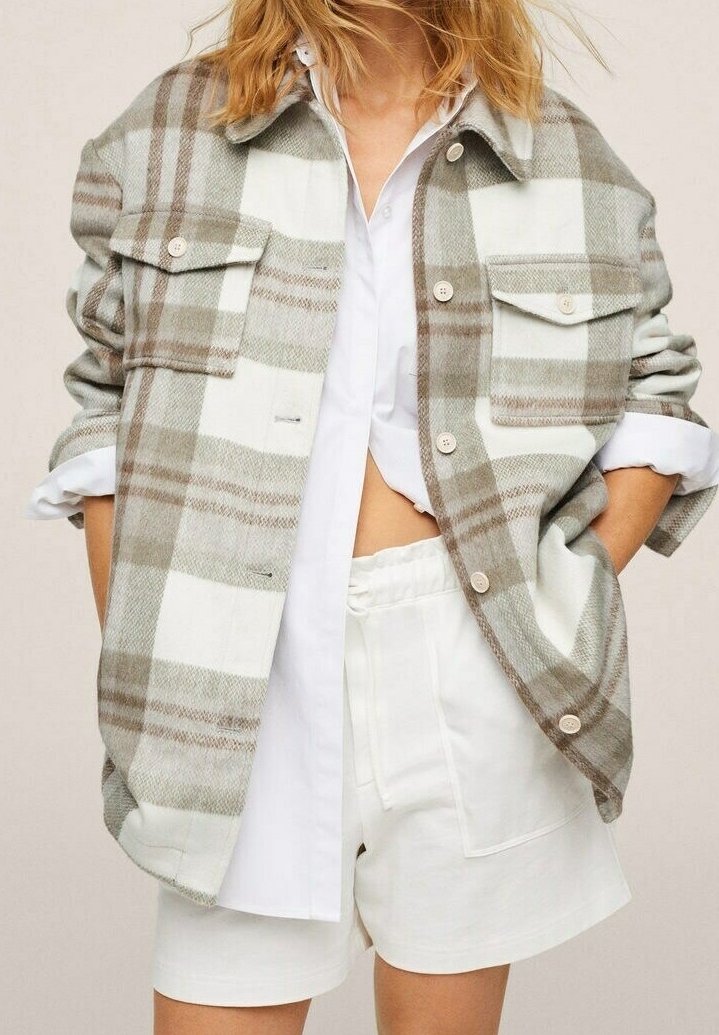 Personne blonde portant une veste à carreaux beige et blanche boutonnée par-dessus une chemise blanche et un short blanc à taille élastique, les mains dans les poches.