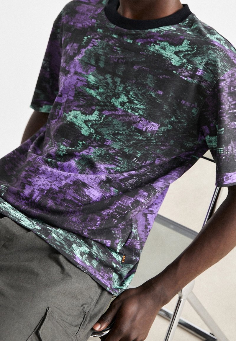 T-shirt à manches courtes noir avec un motif abstrait violet et vert, doté d'un col rond et d'une texture douce.