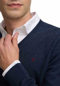 Hombre ajustando el cuello de una camisa blanca que lleva puesta debajo de un suéter de pico azul oscuro con un pequeño logotipo rojo bordado en el pecho.