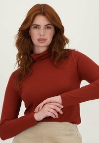 Langarm, roestkleurig shirt met een hoge hals, gemaakt van zachte stof. Het model heeft golvend haar en staat ontspannen met gekruiste handen.