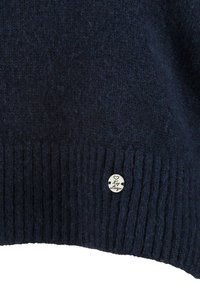 Dunkelblau gestrickter Pullover mit einem gerippten Saum und einem runden silbernen Etikett mit der Aufschrift "Key Large", das an der unteren linken Seite angebracht ist.
