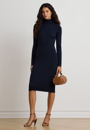 Femme portant une robe midi bleu marine cintrée à col roulé, tenant un petit sac à main rond marron et des sandales à talons hauts couleur beige.