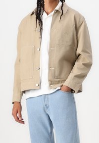 Beżowa bawełniana kurtka z zatrzaskami, dwoma przednimi kieszeniami i małym logo na piersi. Noszona na białej koszuli z jasnoniebieskimi dżinsami.