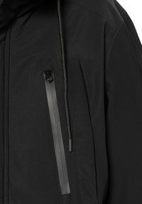 Chaqueta exterior negra con un bolsillo frontal con cremallera, capucha ajustable con cordón y tejido suave con un sutil detalle de costuras.