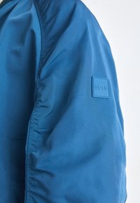 Blå jacka med en jämn textur, med en rynkad ärm och ett fyrkantigt logotypmärke märkt "BOSS" på vänster arm.