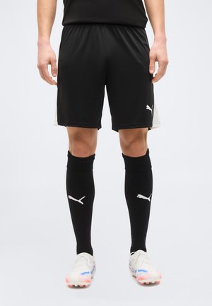 Fekete Puma sportnadrágot, fekete térdig érő Puma zoknit és fehér, piros-kék részletekkel díszített stoplis cipőt viselő személy áll fehér háttér előtt.