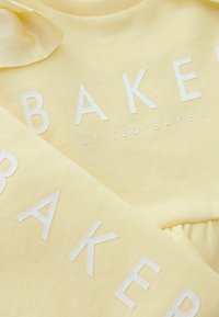Žuti pamučni dukserica s metalik srebrnim natpisom "BAKER" s blagom teksturom, dopunjen nježnom trakicom na izrezu.