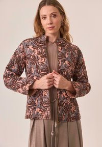 Mujer vistiendo una chaqueta acolchada con un patrón de hojas marrones y rosas, un top de punto topo y una falda topo, ajustando la cremallera de la chaqueta.