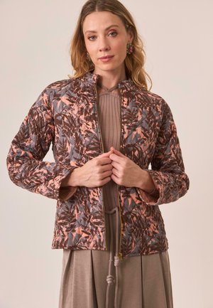 Mujer vistiendo una chaqueta acolchada con un patrón de hojas marrones y rosas, un top de punto topo y una falda topo, ajustando la cremallera de la chaqueta.