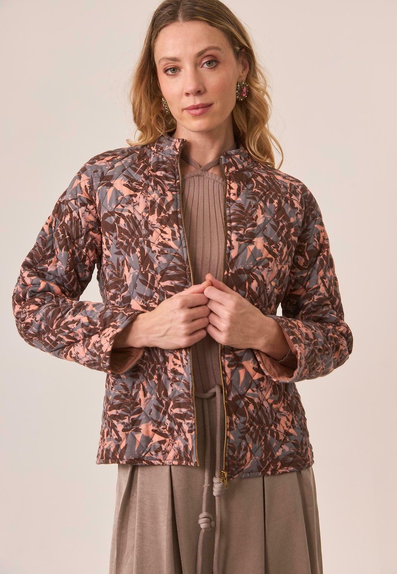 Mujer vistiendo una chaqueta acolchada con un patrón de hojas marrones y rosas, un top de punto topo y una falda topo, ajustando la cremallera de la chaqueta.