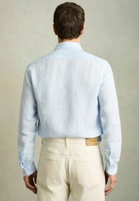 Camisa de lino azul claro de manga larga con cuello clásico y puños abotonados, combinada con pantalones de color crema con bolsillos traseros y etiqueta de logo.