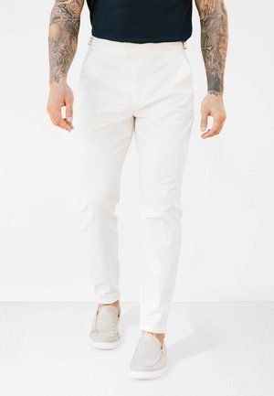 Pantaloni albi, croială slim, cu o textură fină, având fermoare laterale și purtați cu pantofi slip-on gri deschis.