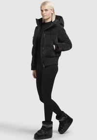 Zwarte pufferjack met een capuchon, rits aan de voorkant en elastische manchetten, gecombineerd met strakke leggings en zwarte laarzen met bontvoering. Glad textuur.