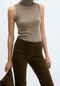 Vaaleanruskea korkeakauluksinen hihaton paita yhdistettynä ruskeisiin corduroihousuihin, joissa on pehmeä pinta ja istuva muotoilu, yllä samaan sarjaan kuuluva takki.