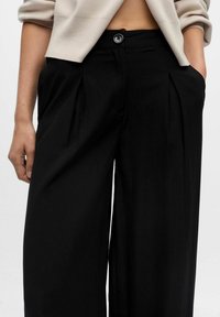 Donna che indossa pantaloni neri a vita alta e gamba larga con pieghe frontali e un maglione beige corto, con le mani nelle tasche.