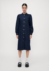 Temno denim obleka z visokim ovratnikom, napihnjenimi dolgimi rokavi, zapiranjem na gumbe in dolžino nad koleni. Ima vidne šive in dva stranska žepa.