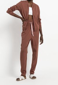 Brun bomullsmixad träningsoverall med zipjacka, långa ärmar och byxor med elastisk midja som har en rosa dragsko. Vita sandaler med remmar.