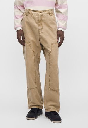 Person iført løse beige bukser med store forlommer og sorte sneakers, stående mod en ensfarvet lys baggrund.