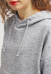 Sudadera gris claro con un tejido texturizado. Presenta una capucha con cordón, un sutil patrón jaspeado y detalles acanalados en el dobladillo y los puños.
