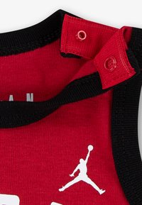 Rød ærmeløs skjorte med sort kant, der har en hvid grafik af en springende figur og trykknapper på skulderen. Fremstillet af bomuldsstof.