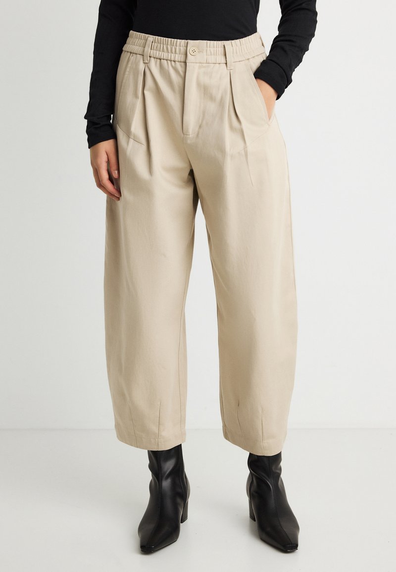 Μπεζ παντελόνι ψηλόμεσο cropped με πιέτες, πλαϊνές τσέπες και κούμπωμα με κουμπί, συνδυασμένο με μαύρο μακρυμάνικο τοπ και μαύρα μποτάκια με τακούνι.