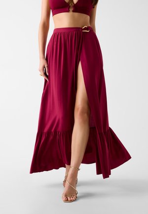 Kobieta ubrana w burgundową krótki top i pasującą maxi spódnicę z wysokim rozcięciem, ozdobioną złotymi klamrami oraz złote sandały na wysokim obcasie z paskami.