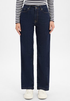 Dunkelblaue weit geschnittene Jeans mit hoher Taille, ausgestattet mit orangefarbenen Nähten, fünf Taschen und einem klassischen Knopfverschluss. Kombiniert mit weißen Turnschuhen.