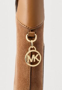 Bolso de ante marrón vista lateral con una parte superior de cuero liso, que presenta una etiqueta de logo circular dorada con "MK" adjunta a una correa de cuero.