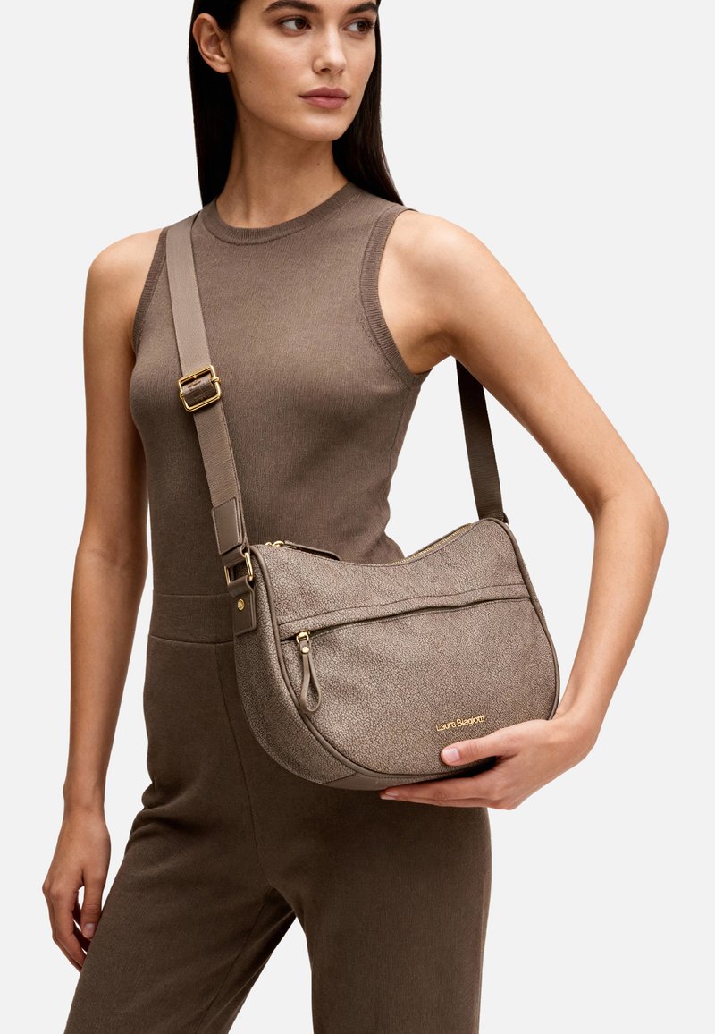 Donna con outfit marrone senza maniche tiene una borsa a tracolla taupe con texture, cerniera dorata e logo "Laura Biagiotti".