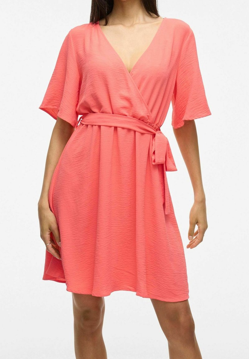 Korálově oranžové zavinovací šaty z lehké texturované látky. Mají krátké rukávy, hluboký výstřih do V a detail vázání v pase. Volný splývavý střih.
