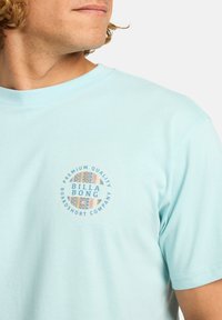 T-shirt in cotone azzurro chiaro con un logo grafico circolare che riporta la scritta "BILLA BONG" e motivi geometrici sul petto. Design con scollatura a girocollo.