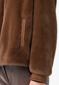 Dettaglio ravvicinato della manica e della tasca di una giacca in finta pelliccia marrone, abbinata a pantaloni texturizzati marrone scuro.