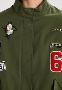 Oliwkowa kurtka z wysokim kołnierzem, ozdobiona haftowanymi różami, czerwonym "6" oraz metalowymi ćwiekami na kieszeniach. Teksturowana tkanina z łatami.