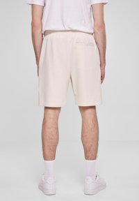 Homme portant un short couleur crème et des baskets blanches, se tenant de dos face à l'objectif contre un fond gris uni.