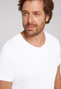 White cotton t-shirt with a crew neck, short sleeves, and a smooth texture. Model features light stubble and shoulder-length wavy hair.