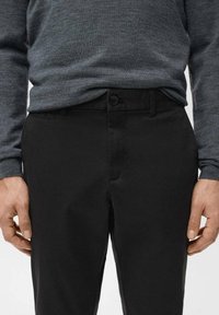 Mężczyzna ubrany w ciemnoszarą koszulę z długim rękawem włożoną do czarnych spodni z zapięciem na guziki i zamek, ręce swobodnie opuszczone wzdłuż ciała.