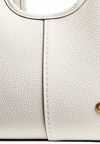 Gros plan sur un cuir grainé blanc avec une couture centrale et une partie d’un bouton en laiton au bord d’un sac ou accessoire.